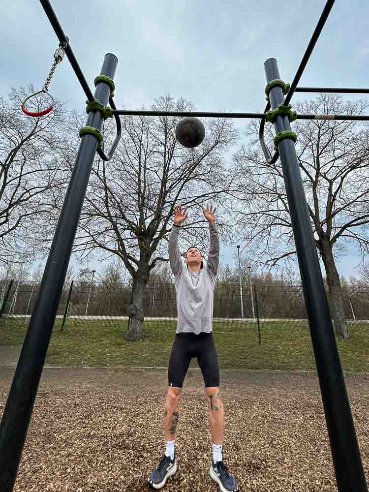 Ein Sportler wirft in einem Outdoor-Trainingspark einen Medizinball nach oben unter ein Calisthenics-Gerüst. Die Szene zeigt eine funktionelle Kraft- und Ausdauerübung, wie sie sich gut für HYROX-Training ohne Gym eignet.