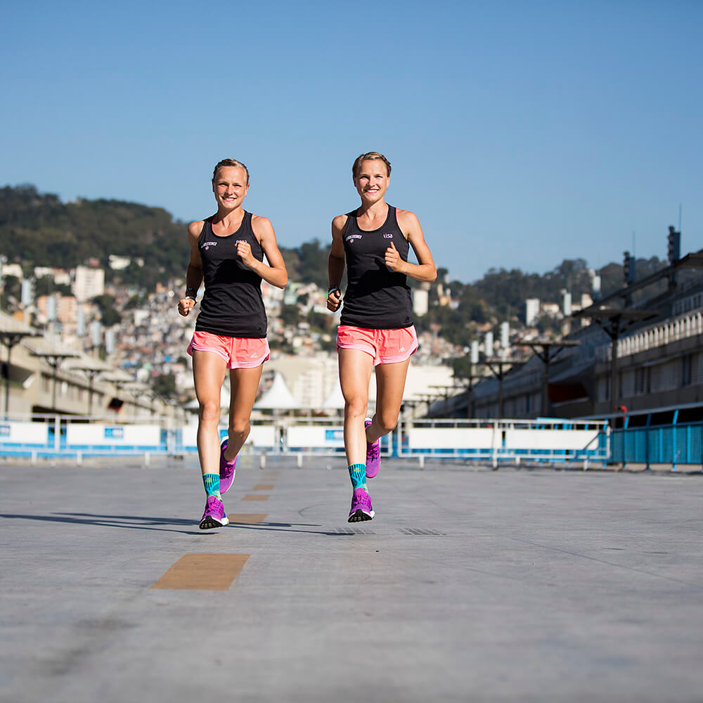 Sprunggelenkbandage beim Laufen Hahner Twins laufen auf einer Straße lächelnd auf die Kamera zu	 die tragen Sprunggelenkbandagen für den Sport. Im Hintergrund ist eine Stadt und Hügel zu sehen.