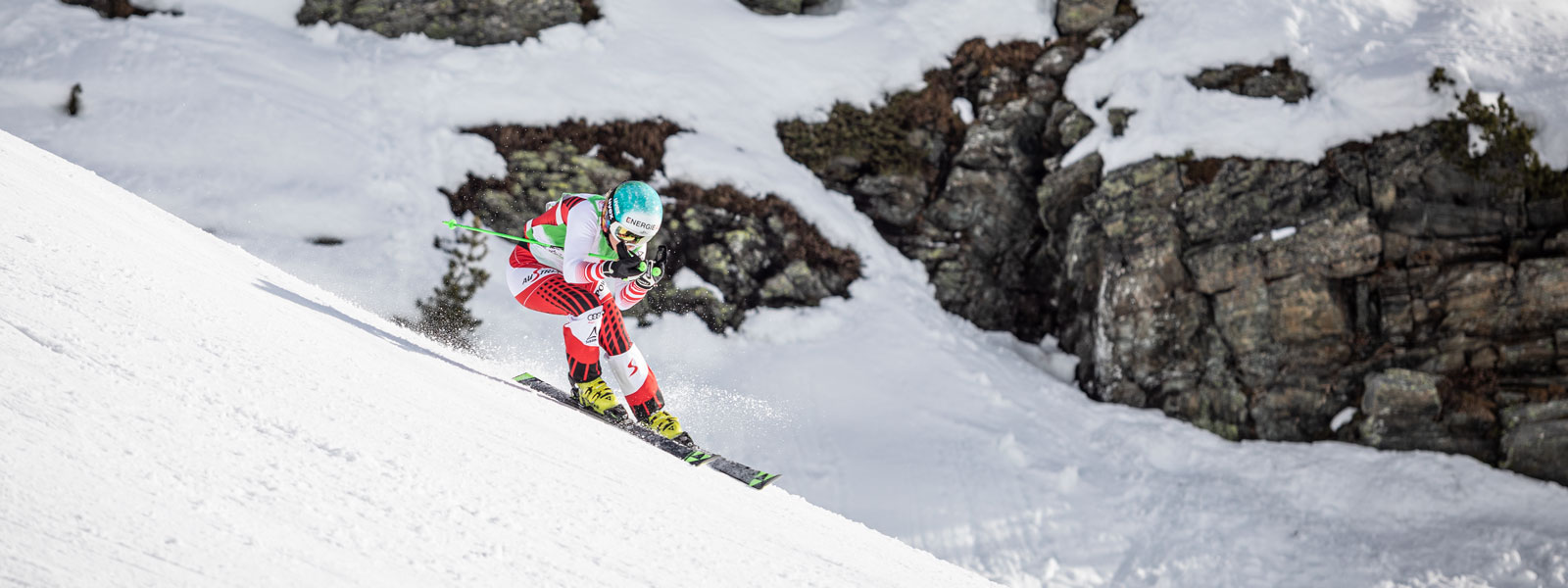 Ab in die Abfahrtshocke: Genieße Deinen Winter auf der Piste und mit unseren Produkten Skiläuferin im rot-weißen Rennanzug in Abfahrtsposition bei einem leichten Schwung mit großen	 schneebedeckten Felsen im Hintergrund
