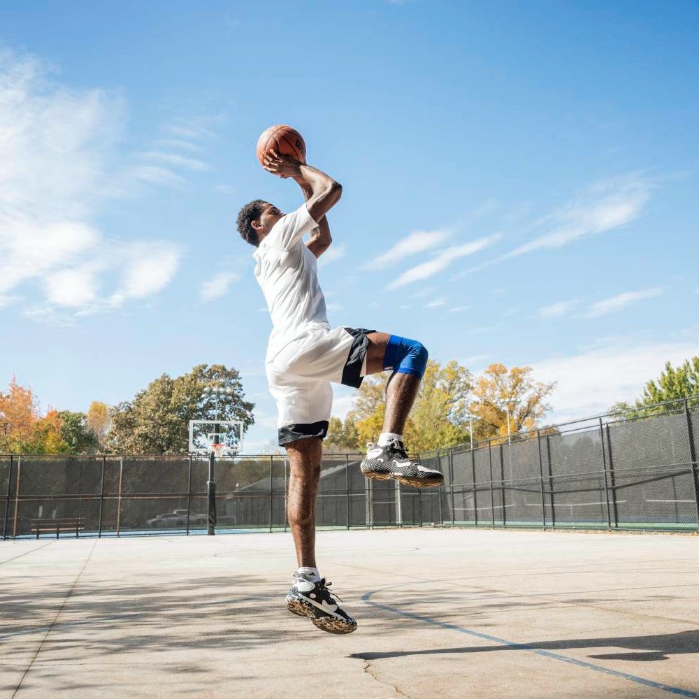 Dallas Mavericks Support – Höher springen, sicher landen Ein Basketballspieler mit einer blauen Dallas Mavericks-Kniebandage beim Sprungwurf auf einem Outdoor-Court. Die Bandage unterstützt das Knie während der Bewegung.