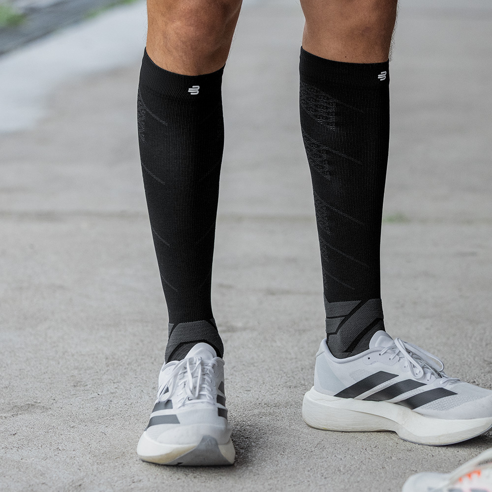 Ein sportlicher Mann steht frontal auf Asphalt, bekleidet mit schwarzen Kompressionsstrümpfen von Bauerfeind und weißen Laufschuhen. Die geradlinige Haltung und die Muskelspannung in den Waden betonen den stabilisierenden Effekt der Socken im Stehen.