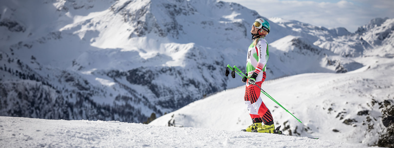 Skifahrerin im rot-weißen Rennanzug steht lächelnd und mit den Stöcken in den Händen vor einem gewaltigen Bergmassiv