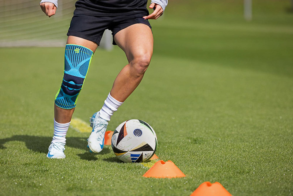 Eine Fußballspielerin trainiert auf dem Platz und trägt eine Bauerfeind Kniebandage. Der Fokus liegt auf der Stabilität und Unterstützung, die die Bandage während dynamischer Bewegungen bietet, wie das Dribbeln um Hütchen.