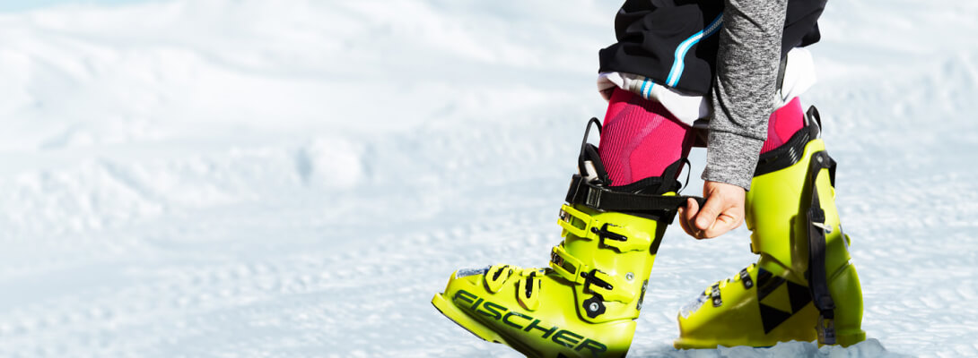 Bildauschnitt zeigt eine Person in Skistiefel mit Skisocken darunter	 sie greift an den Verschluss der Stiefel