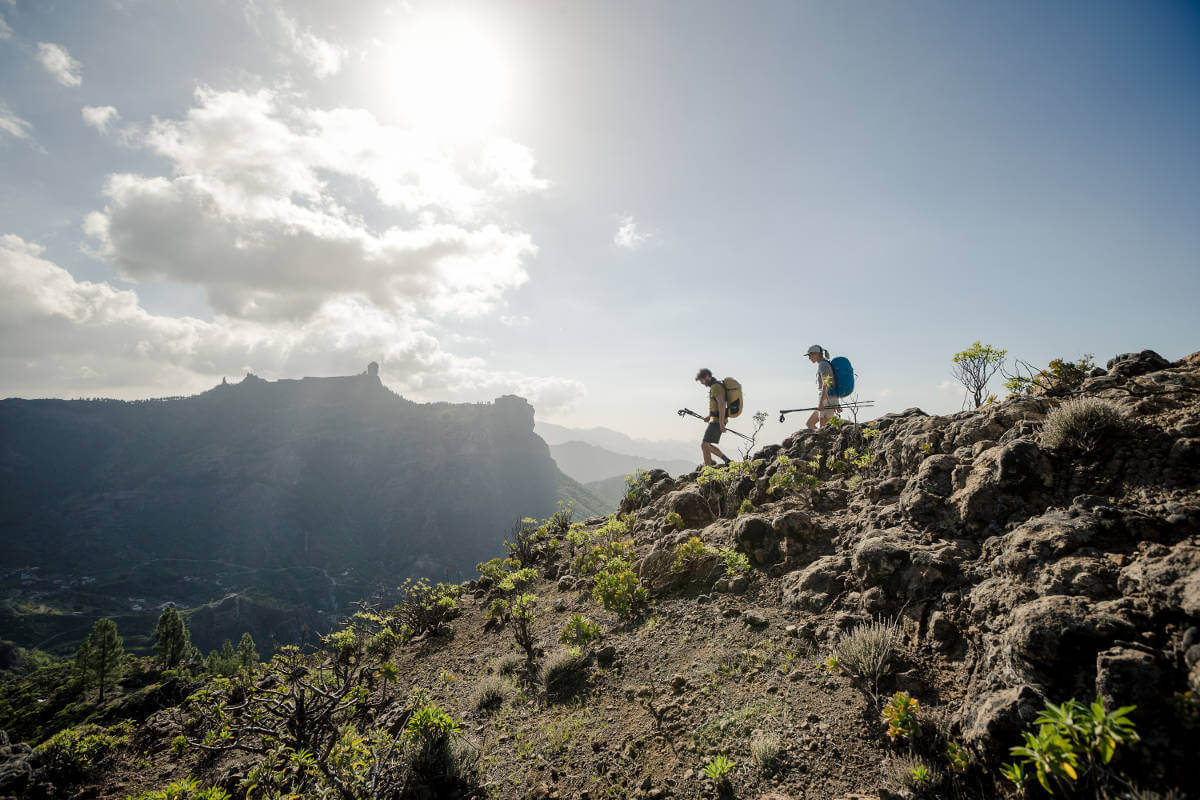 Weitwinkel: Zwei Wanderer mit großen Rucksäcken und Stöcken gehen vorsichtig einen Gebirgsweg mit Steinen und kleinen Sträuchern nach unten	 im Hintergrund sind weitere Berge zu sehen