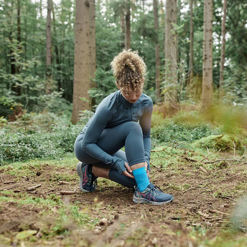 Eine Läuferin kniet auf dem Boden im Wald und richtet sich ihre Achilles Bandage