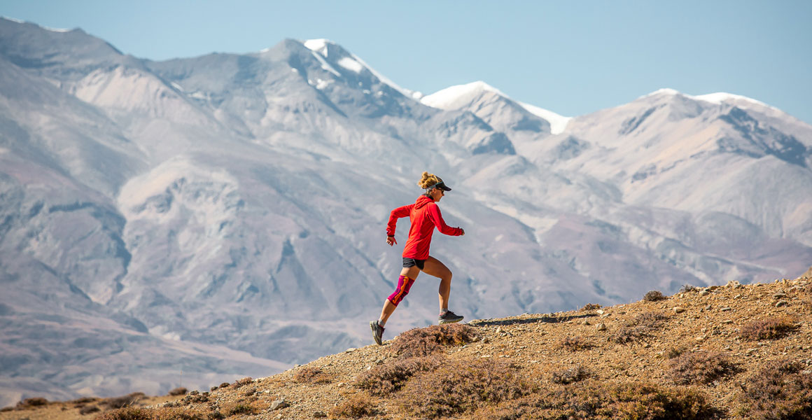 Trailläuferin läuft mit dem Himalaya im Hintergrund einen leichten Anstieg hinauf und trägt eine Kniebandage