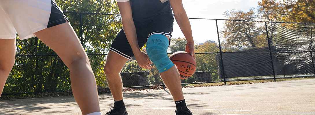 Knee Sleeve beim Basketball Ein Basketballspieler steht auf einem betonierten Platz der umzeunt ist und macht ein Trick mit dem Ball bei dem er durch seine beiden Beine spielt. Ihm gegenüber steht ein weiterer Mitspieler. Er trägt dabei ein Knee Sleeve.
