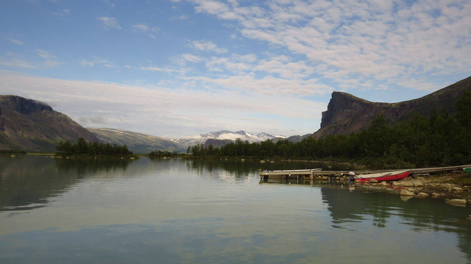 Panorama eines Sees umgeben von Bäumen und kleinen Bergen	 am rechten Rand liegt ein kleines Boot am Steg