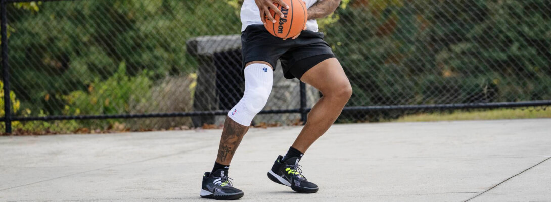 Spieler mit weißer Kniebandage beim Ballhandling Basketballspieler mit weißer Kompressions-Kniebandage beim Ballhandling auf einem Outdoor-Court. Die weiße Bandage kontrastiert mit der dunklen Haut und der schwarzen Shorts und trägt ein gut sichtbares NBA-Logo.