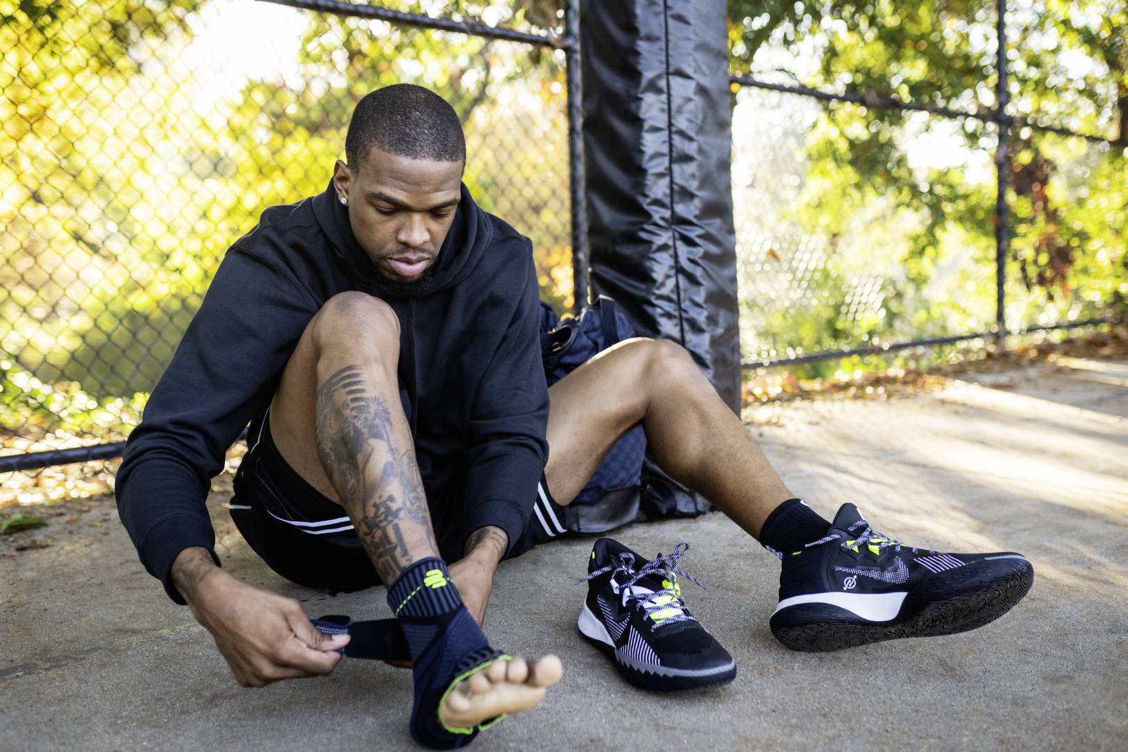 Basketballspieler mit Hoodie zieht sich am Rand eines Streetball-Courts eine Sprunggelenkbandage mit Gurt an