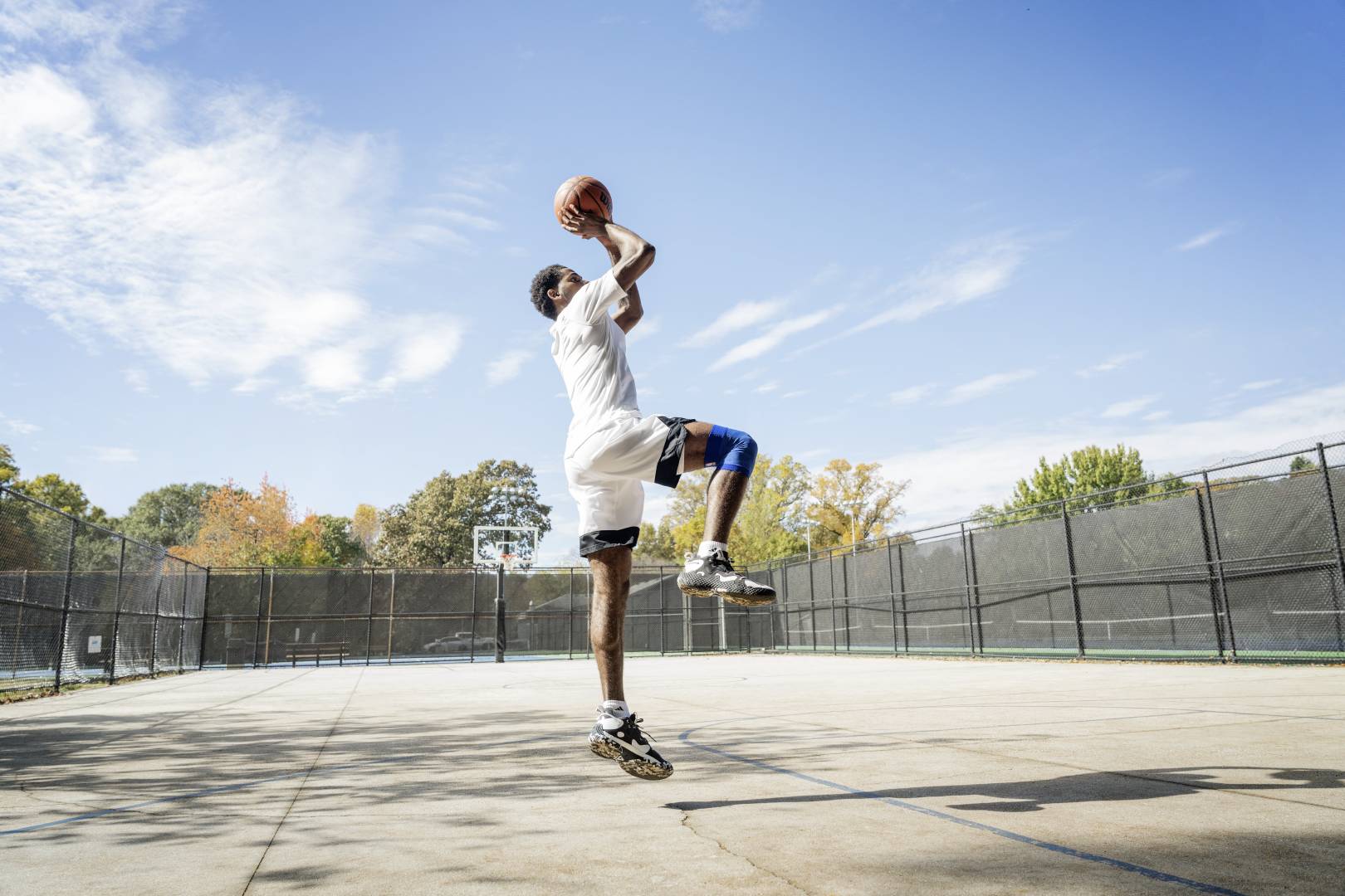 Basketballer springt auf einem Streetcourt für den Wurf ab	 Hände mit Ball überm Kopf	 ein Bein angewinkelt	 das andere senkrecht nach unten zeigend