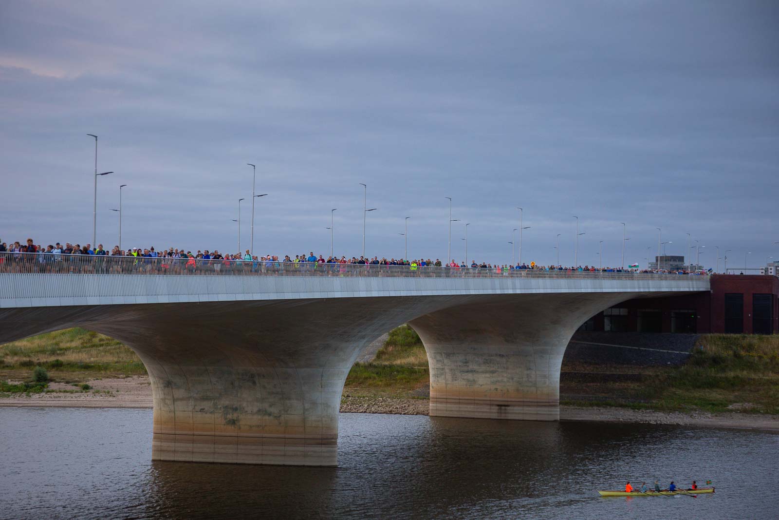 Zahlreiche Teilnehmer des 4Daagse marschieren im Morgengrauen über eine breite Brücke in Nijmegen, während unten ein Ruderboot vorbeifährt.